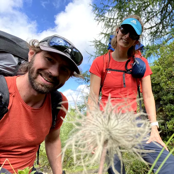 Twee wandelaars in rode shirts en met een zonnebril op glimlachen naar de camera in een zonnige, groene buitenomgeving, met een pluizige witte bloem op de voorgrond en een blauwe lucht met wolken achter hen.